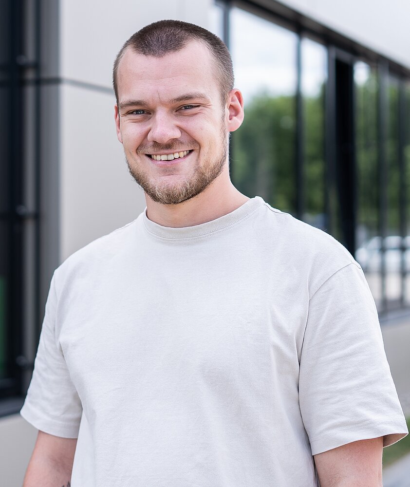 Noah Hilbrecht stands smiling in front of the camera, the AfB building can be seen behind him. He is wearing a beige T-shirt.