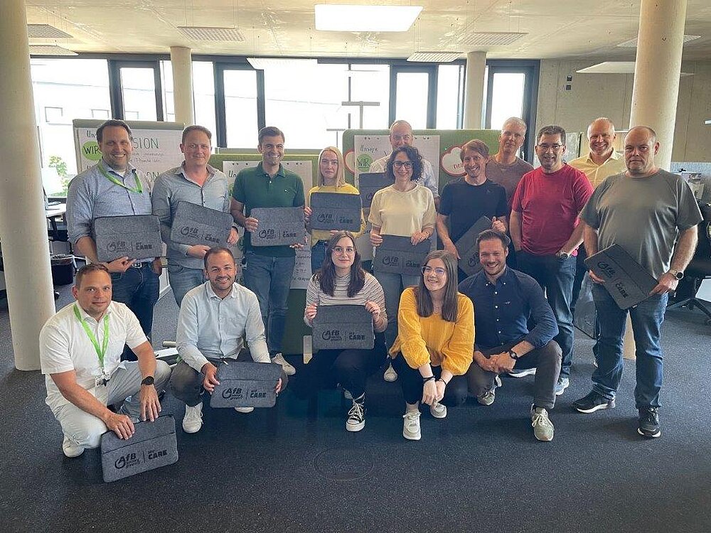 Eine Gruppe von 16 Mitarbeitenden bei AfB macht ein Teamfoto. Sie lachen und halten ihren Laptophüllen mit einem "CARE"-Motiv in die Kamera.