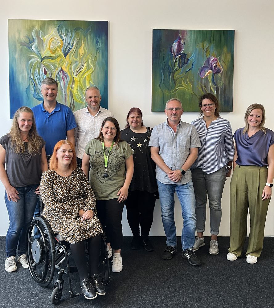 A group of nine people smile cheerfully into the camera. They are posing in front of two abstract flower paintings in shades of green and blue. They are all standing, except for one person. Marina Zdravkovic is sitting in a wheelchair. Pictured from top left to bottom right: Andreas Melzer, Sven Mersi, Tanja Schmettlach, Werner Ahles, Christina Gebhard, Yvonne Cvilak, Anja Spangenberg, Marina Zdravkovic and Yasmin Stößer.