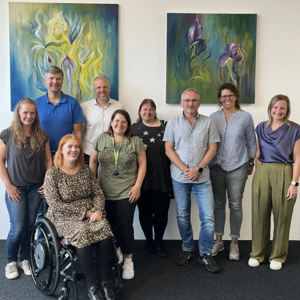 A group of nine people smile cheerfully into the camera. They are posing in front of two abstract flower paintings in shades of green and blue. They are all standing, except for one person. Marina Zdravkovic is sitting in a wheelchair. Pictured from top left to bottom right: Andreas Melzer, Sven Mersi, Tanja Schmettlach, Werner Ahles, Christina Gebhard, Yvonne Cvilak, Anja Spangenberg, Marina Zdravkovic and Yasmin Stößer.
